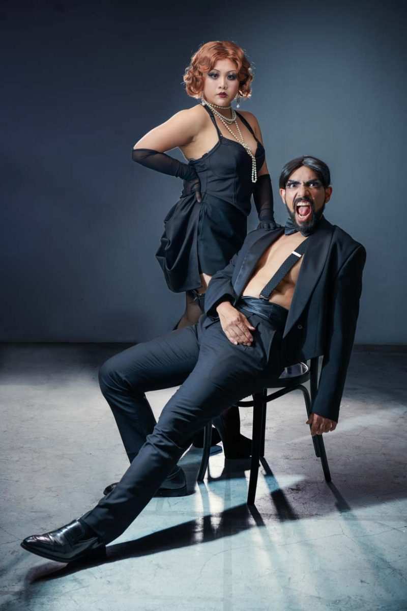 Singapore advertising portrait photography: publicity portrait. A woman in a black corset dress, pearls and gloves standing behind a man in a black suit seated on a chair, studio portrait for Sing'Theatre's Cabaret