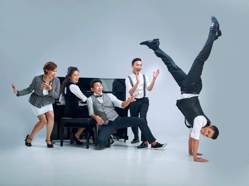 Singapore advertising portrait photography: theatre key art. The cast of A Spoonful of Sherman around a piano in a white studio, one performer frozen in a handstand, group portrait for Sing'Theatre's key visual
