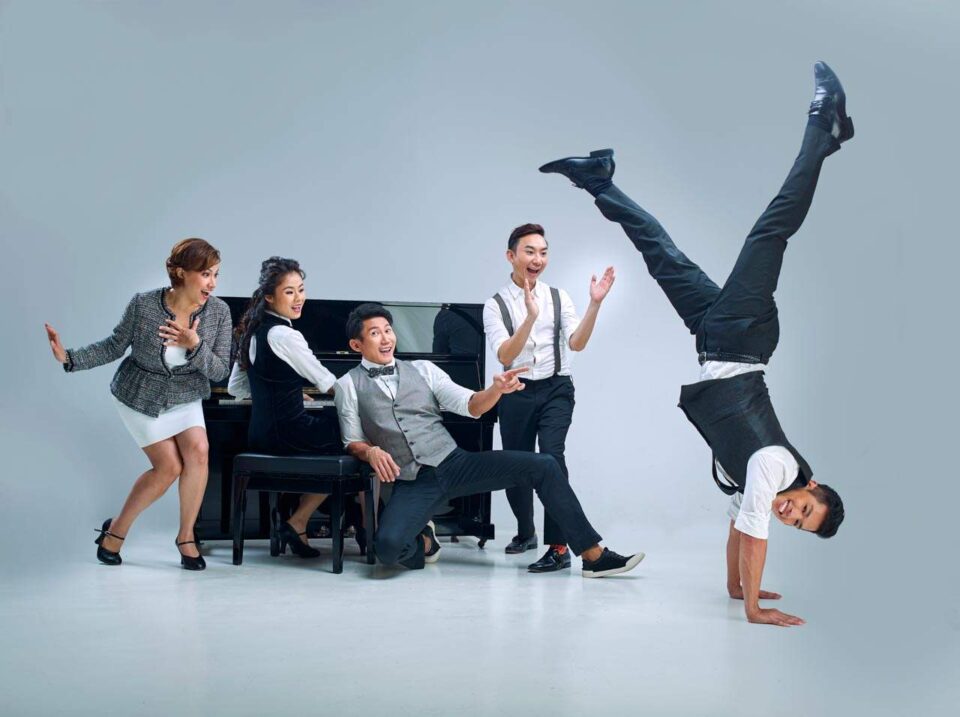 Singapore advertising portrait photography: theatre key art. The cast of A Spoonful of Sherman around a piano in a white studio, one performer frozen in a handstand, group portrait for Sing'Theatre's key visual