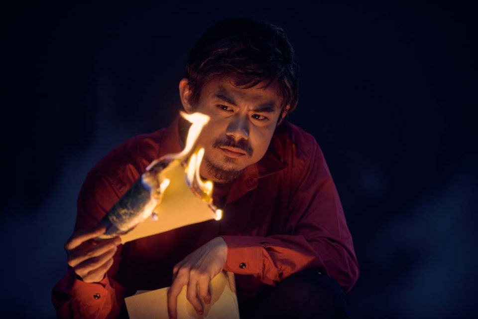 Singapore advertising portrait photography: theatre key art. A performer lifting a burning sheet of paper to his face in the dark, production visual for The Theatre Practice's 四马路