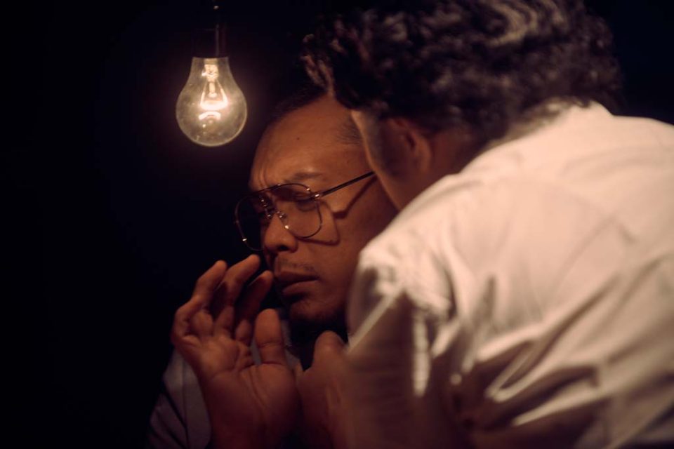 Singapore advertising portrait photography: theatre key art. A man in glasses beneath a bare light bulb, a hand raised to his face beside another figure's shoulder, production visual for The Theatre Practice's 四马路