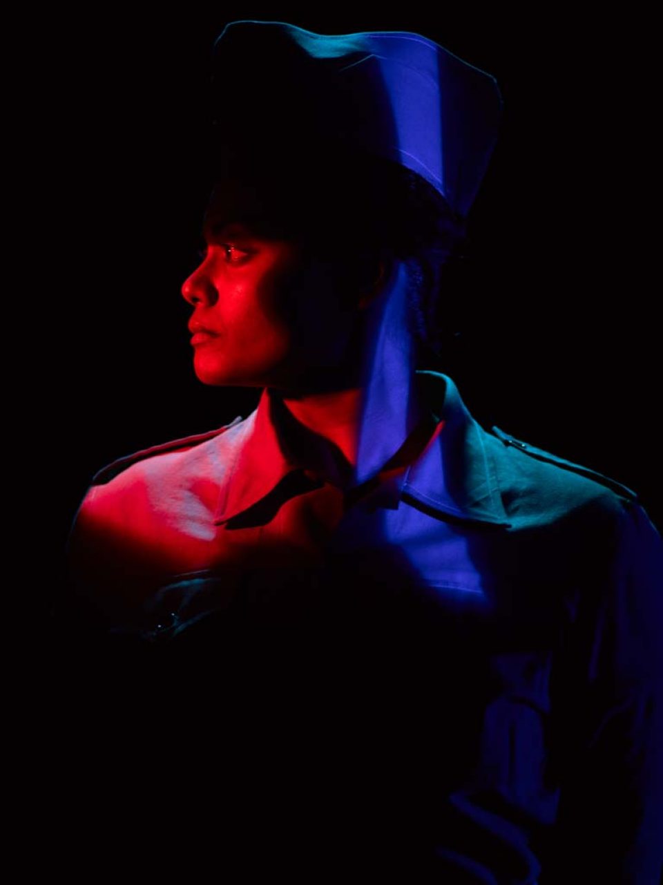 Singapore advertising portrait photography: theatre key art. A performer in a peaked cap in profile, face and shoulder cut by red and blue light against black, studio production visual for theatre publicity