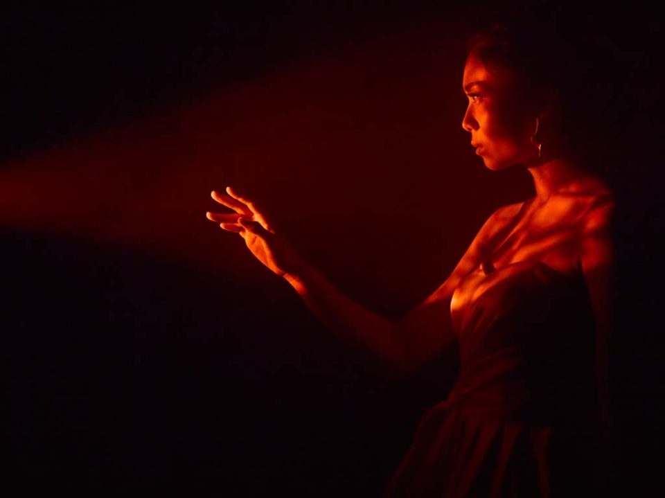 Singapore advertising portrait photography: theatre key art. A woman in a dark dress reaching into a beam of warm orange light in the dark, studio key visual for a theatre performance