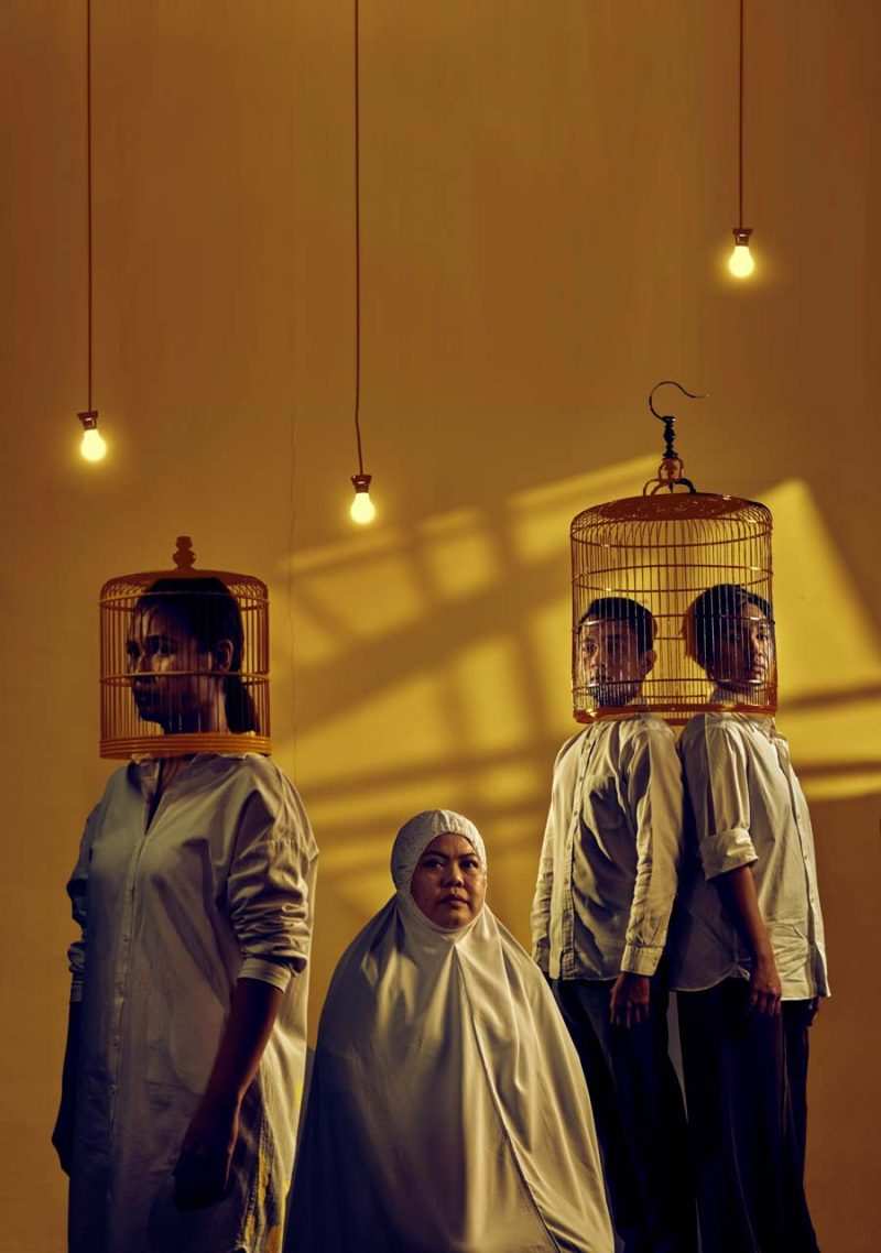 Singapore theatre photography: production photography. Four performers of Oo-Woo in warm window light under hanging bulbs, two with birdcages over their heads, one in a white headscarf and robe, for The Necessary Stage