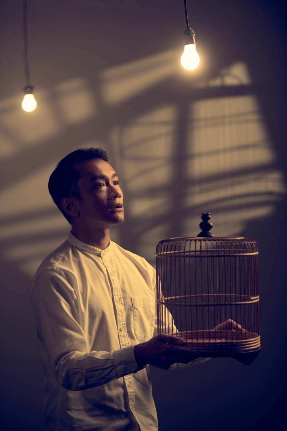 Singapore advertising portrait photography: publicity portrait. A performer in a white shirt holding an empty birdcage in slatted window light under bare bulbs, studio portrait for The Necessary Stage's Oo-Woo