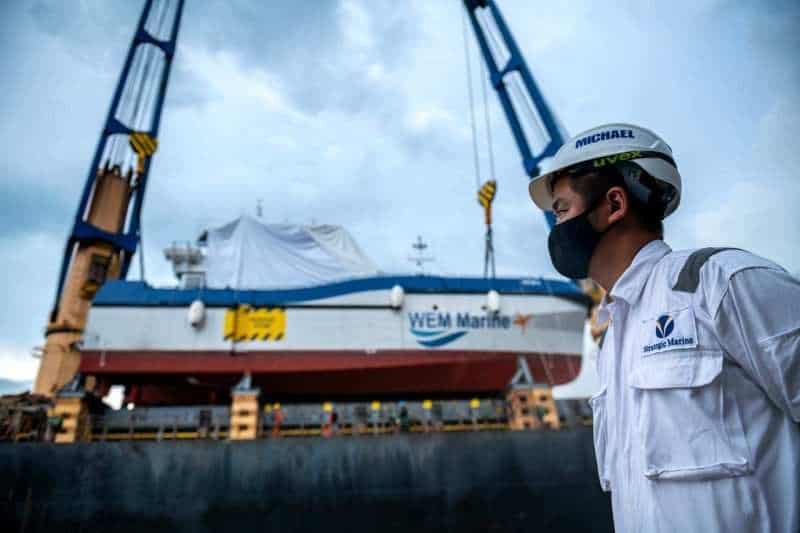 Marine engineer in a white hard hat watching a crawler crane lift a shrink-wrapped WEM Marine unit on the quayside — Singapore industrial photographer