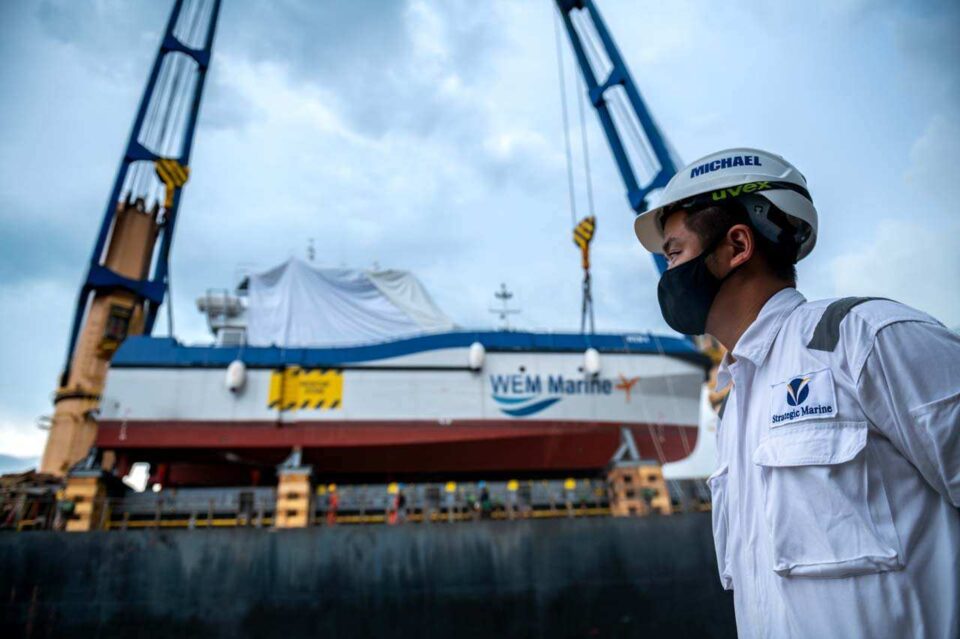 Marine engineer in a white hard hat watching a crawler crane lift a shrink-wrapped WEM Marine unit on the quayside — Singapore industrial photographer