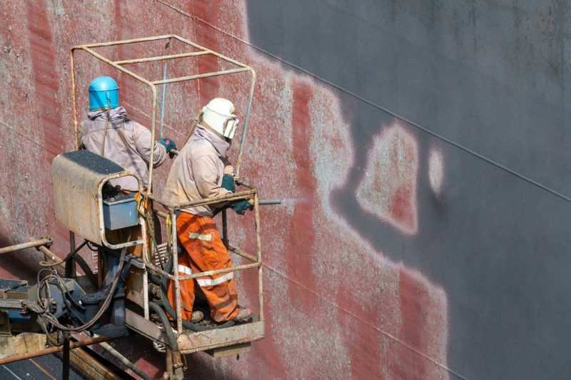 Two yard workers in orange coveralls spray painting a vessel hull from a suspended cradle — Singapore industrial photography, men at work
