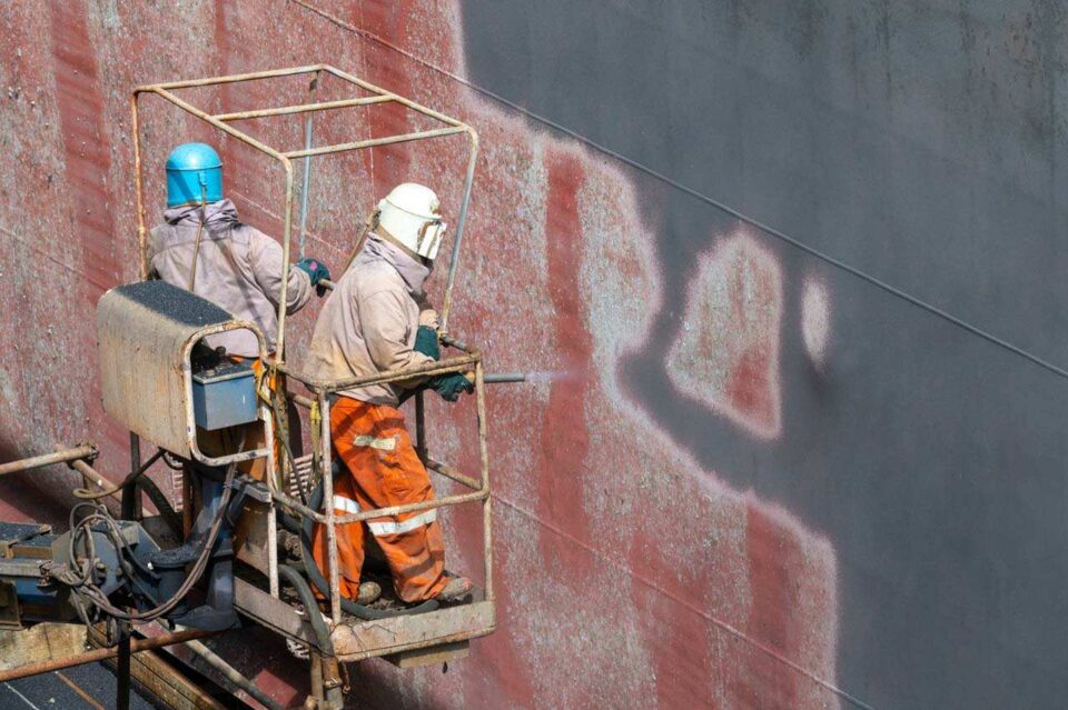 Two yard workers in orange coveralls spray painting a vessel hull from a suspended cradle — Singapore industrial photography, men at work