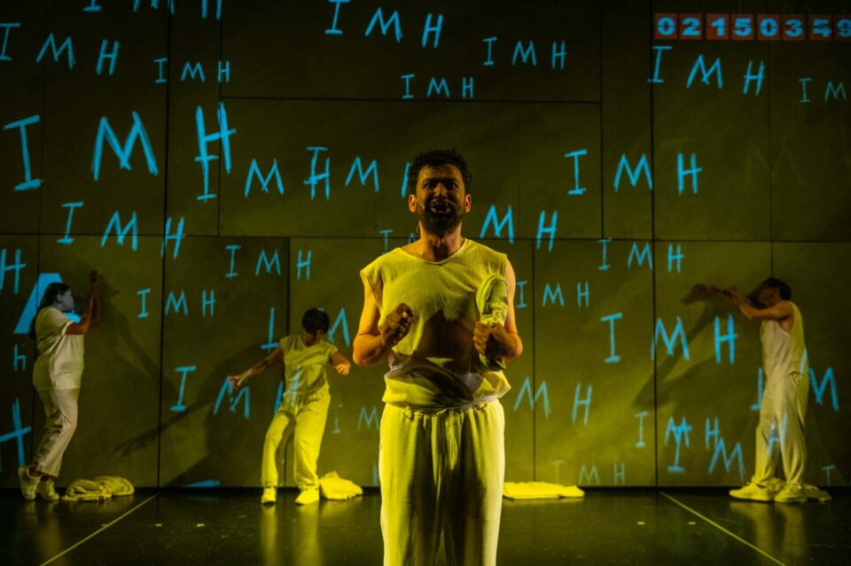 Singapore theatre photography: production photography. A performer in yellow stands centre stage with hands raised as the letters IMH are projected across the walls, in Acting Mad