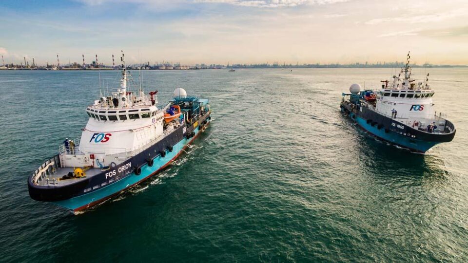Support vessels FOS Orion and FOS Virgo running in close formation off Singapore, industrial shoreline behind — aerial drone photography of vessels in Singapore