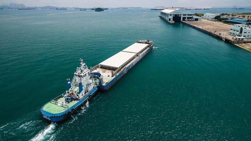 Tug and covered ash barge on the Semakau waste run, approaching the transfer station berth, from the air — aerial drone photography, Singapore