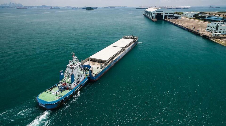 Tug and covered ash barge on the Semakau waste run, approaching the transfer station berth, from the air — aerial drone photography, Singapore