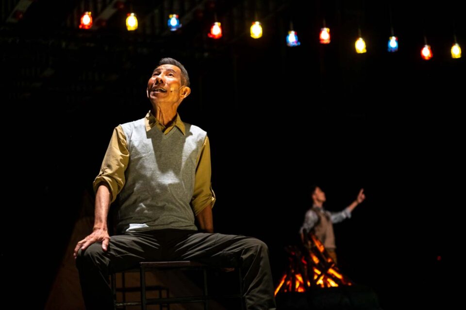 Singapore theatre photography: stage photography. An older man looks up at a string of coloured bulbs while a small figure far behind, beside a fire, gestures upward too, in All The World Is One's Stage