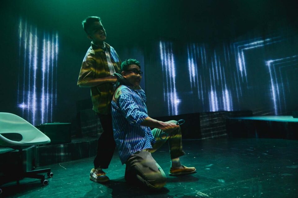 Singapore theatre photography: production photography. In green light one performer stands behind another who kneels, streaks of projected light on the walls, in All The World's A Sea