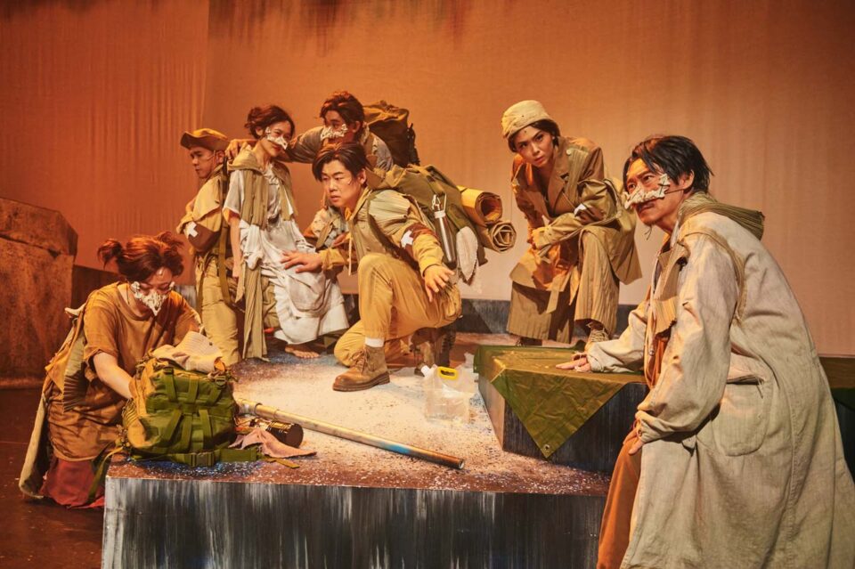 Singapore theatre photography: stage photography. The ensemble in earth-toned costumes crowded on a raised platform in orange light, in All The World's A Sea