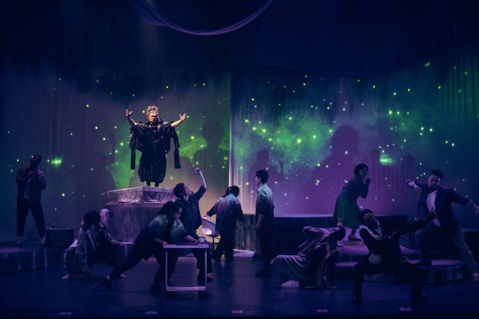 Singapore theatre photography: production photography. The full cast of All The World's A Sea across a dark stage under a projected starfield, one figure raised above them with arms outstretched