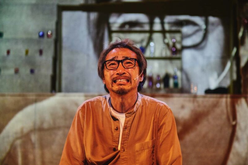 Singapore theatre photography: stage photography. Close-up of an actor in glasses with a strained grin, a projection of his own face looming behind him, in Hi, Can You Hear Me?