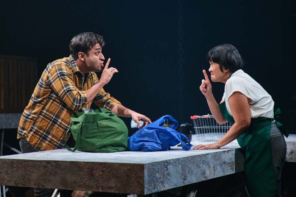 Singapore theatre photography: stage photography. Two actors lean across a table, each with a finger to their lips, shopping bags between them, in No Particular Order