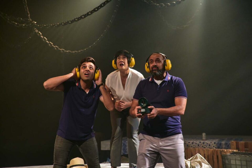 Singapore theatre photography: production photography. Three performers in yellow ear defenders look up in alarm, one holding a device, in No Particular Order