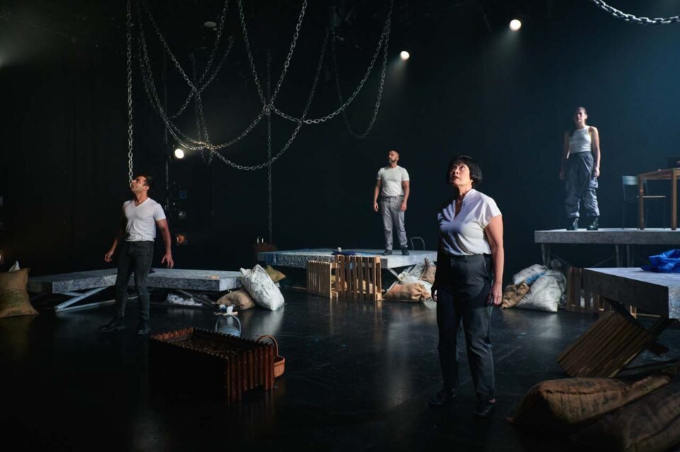 Singapore theatre photography: stage photography. Four performers on a stage hung with chains and strewn with sacks and crates, a woman at centre looking up into the light, in No Particular Order