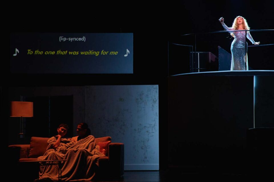 Singapore theatre photography: production photography. A singer in sequins lip-syncs on an upper platform while a couple huddles under a blanket in lamp light below, in Old Gaze