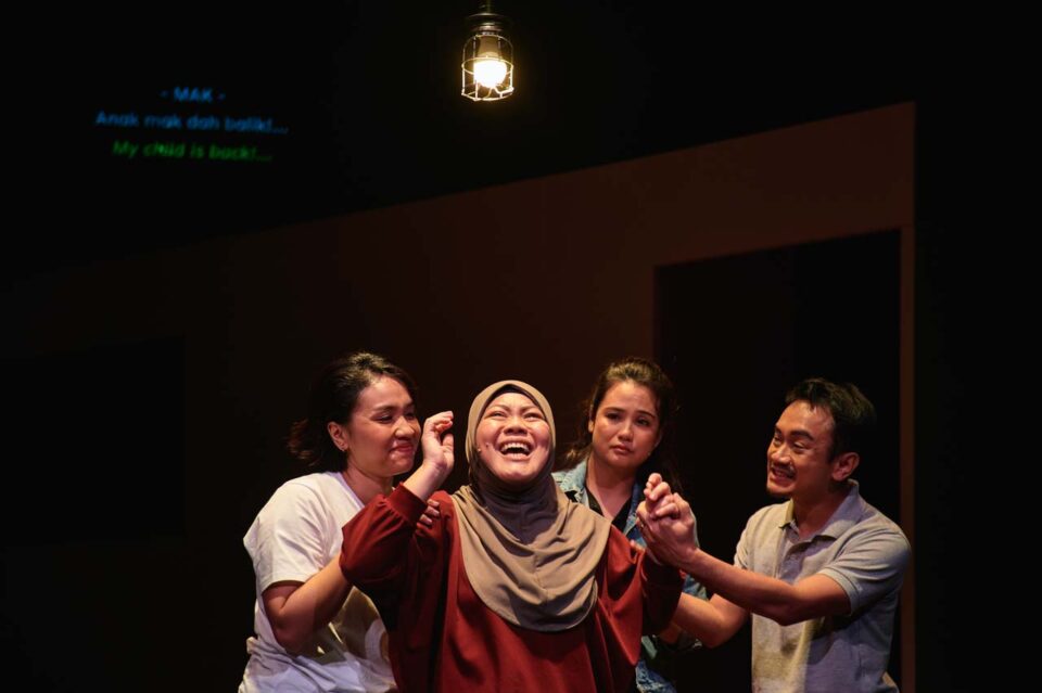 Singapore theatre photography: stage photography. A woman in a hijab bursts into laughter as the three around her react, under the surtitle My child is back!, in Oo-woo