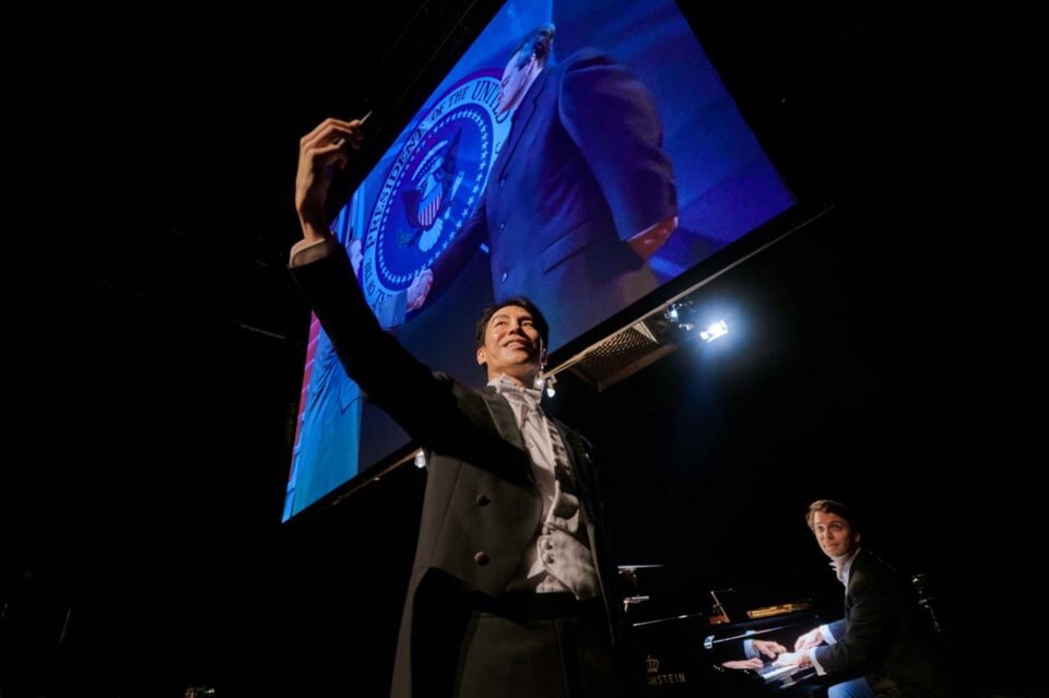 Singapore theatre photography: production photography. A performer in tails raises a phone for a selfie before a projected presidential seal, a pianist playing behind him, in Rhapsody in Yellow