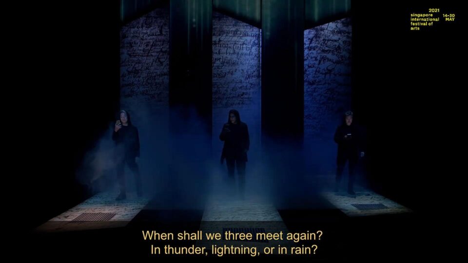 Singapore theatre photography: theatre video still. Video still from the opening of The Commission: three figures in haze between tall walls, subtitled When shall we three meet again?