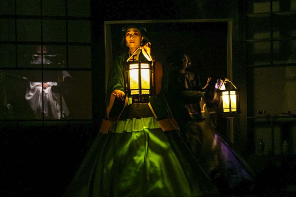 Singapore theatre photography: stage photography. A performer in a green gown carries a lit lantern through the dark as a figure waits in a lit window behind, in The Crab Flower Club