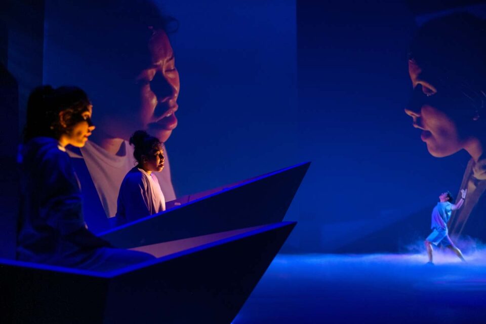 Singapore theatre photography: production photography. Two performers in a giant paper boat beneath huge projected faces, a small figure reaching up through the mist beyond, in The Feelings Farm