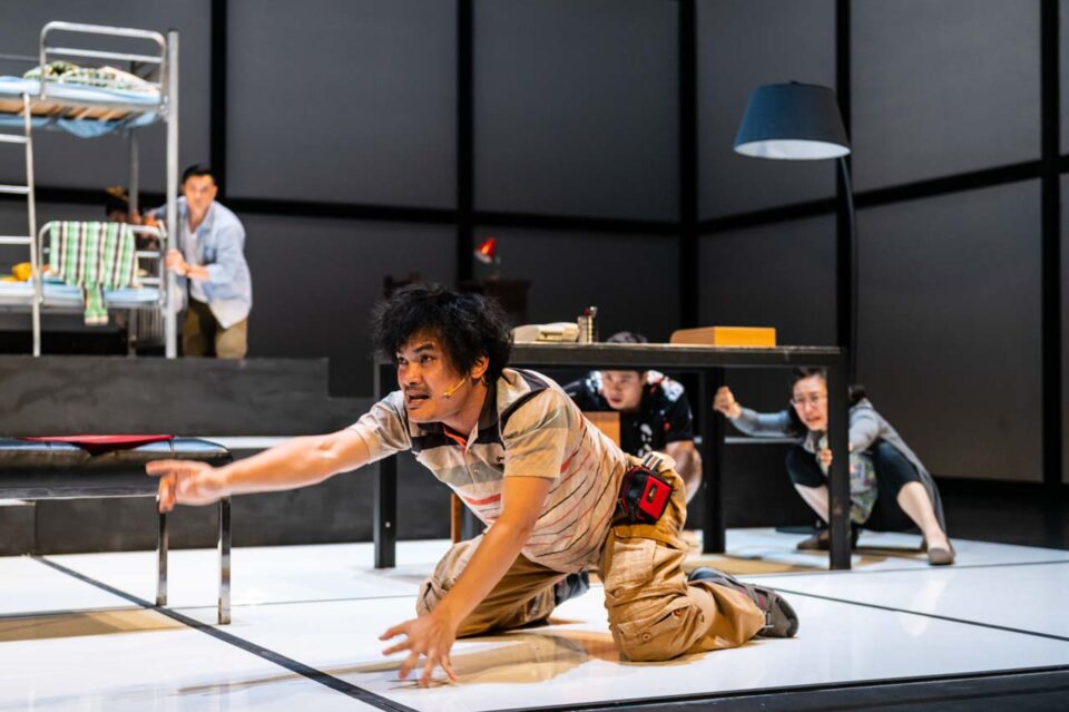 Singapore theatre photography: stage photography. A performer on hands and knees reaches across a bare white floor while others look on from a table behind, in The Way of Zhuang Zi