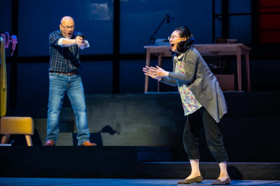 Singapore theatre photography: production photography. A man levels a pistol at a woman in headphones who recoils with her hands up, in The Way of Zhuang Zi